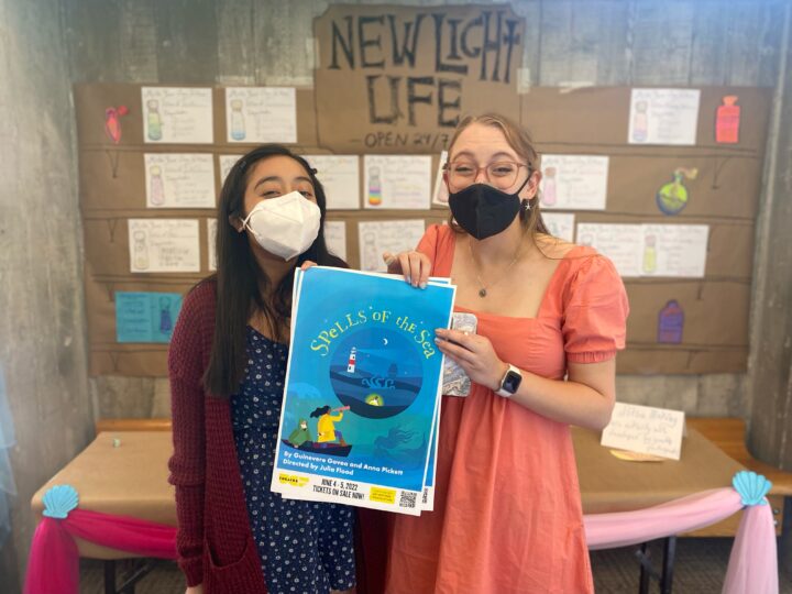 Gwenny Govea and Anna Pickett pose excitedly, holding a poster for their workshop of SPELLS OF THE SEA and standing in front of a cardboard lobby display