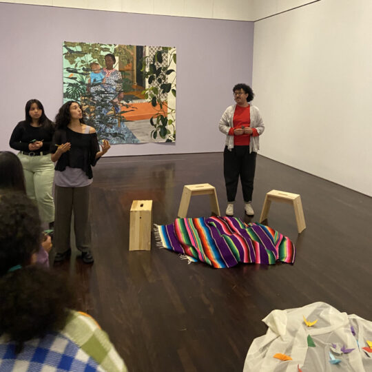 three students stand in front of an audience in a lavender museum gallery with a large, colorful painting behind them and stools in front of them