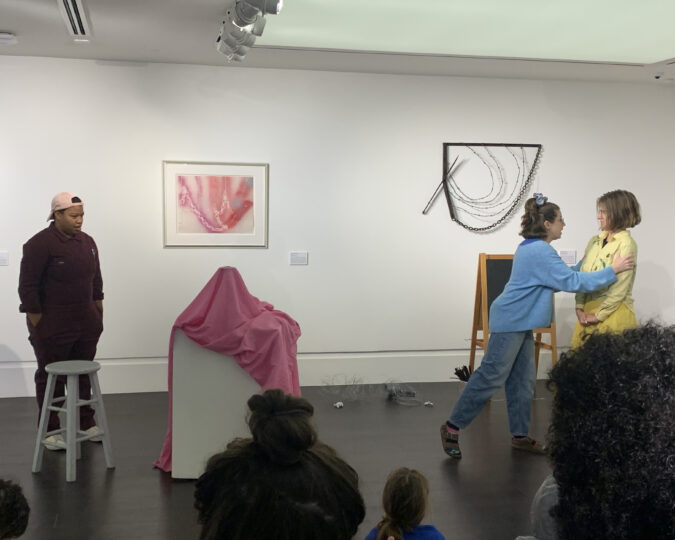 Three actors perform in front of an audience of children, standing in the middle of an art gallery