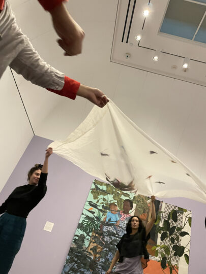 Four students hold up sheer white fabric with colorful origami on top of it, standing in front of a vibrant painting