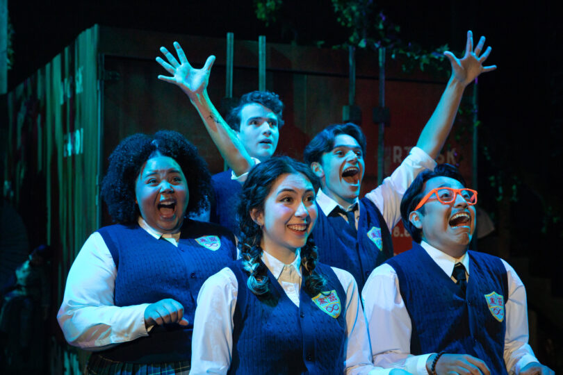 five actors portraying students in a high school choir pose as if sitting on a roller coaster, looking forward with apprehension and excitement