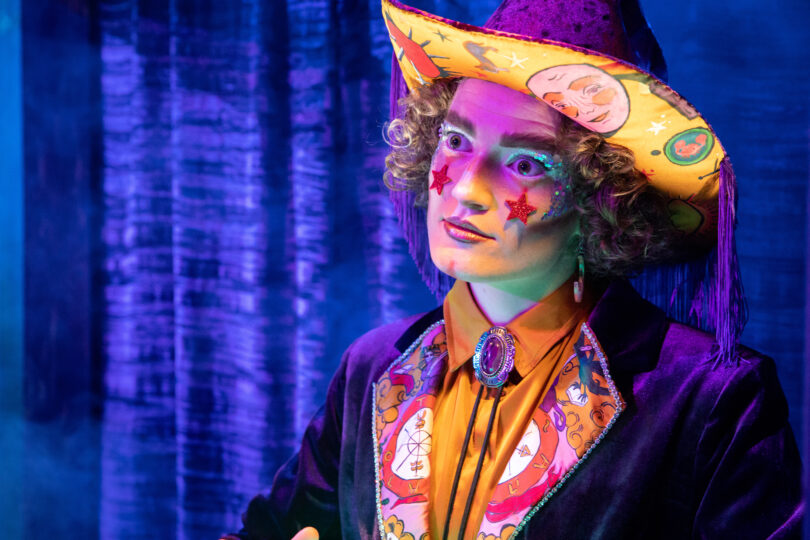 An actor portraying "Karnak," a mechanical fortune teller, stands in front a blue curtain, wearing a velvet purple suit and cowboy hat