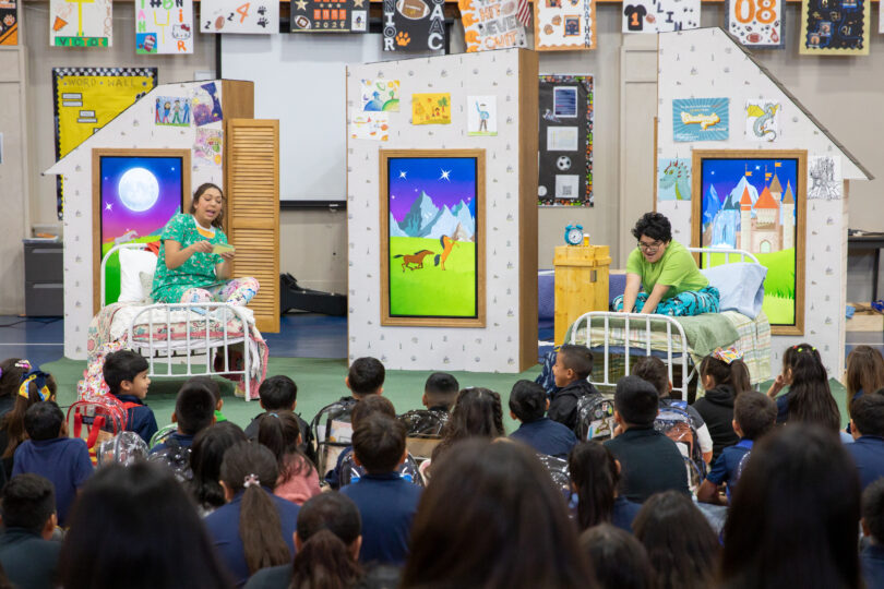 Actors portraying Lizzy and Leo sit on the set for THE SMARTEST GIRL IN THE WORLD in front of an audience of elementary school students