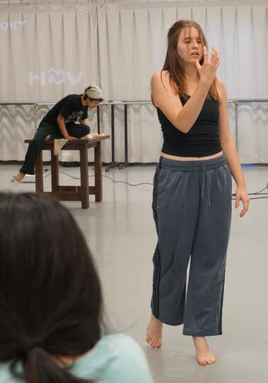 A dancer stands, holding their hand in front of their face, while a choreographer takes notes in the background