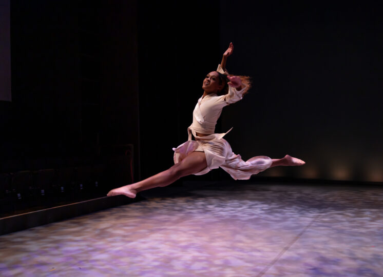 A dancer leaps across stage, their right arm raised above their head and their left arm stretched out beside them