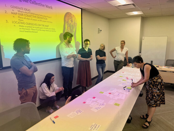 The inaugural group of PT2050 Artist Fellows sit and stand in front of a multicolored projection, listening to Khristián Méndez Aguirre and Katie Dawson share instructions