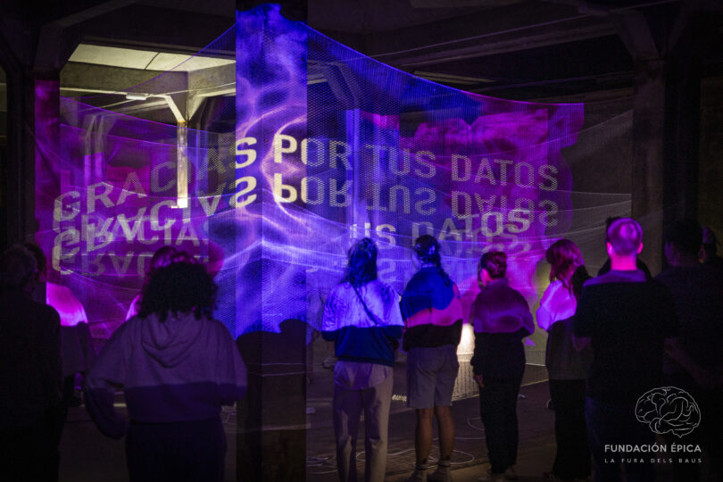 An audiences stands around four columns with a mesh material wrapped around them and vibrant projections reading GRACIAS POR TUS DATOS