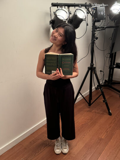 Mia Hsiung Nguyen stands in front of a light tree, holding a book of Anton Chekov's plays open in front of her