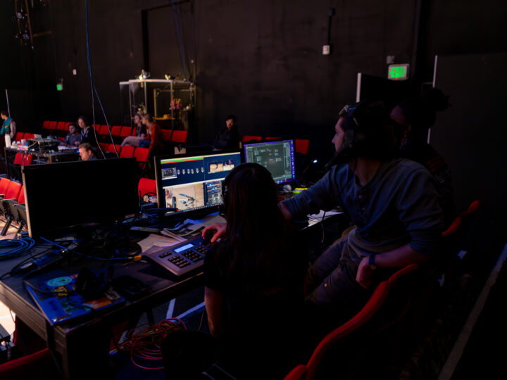 Scenic and IM designer Joshua W. Martin sits at a table in the middle of a seating bank in the theatre, hard at work.