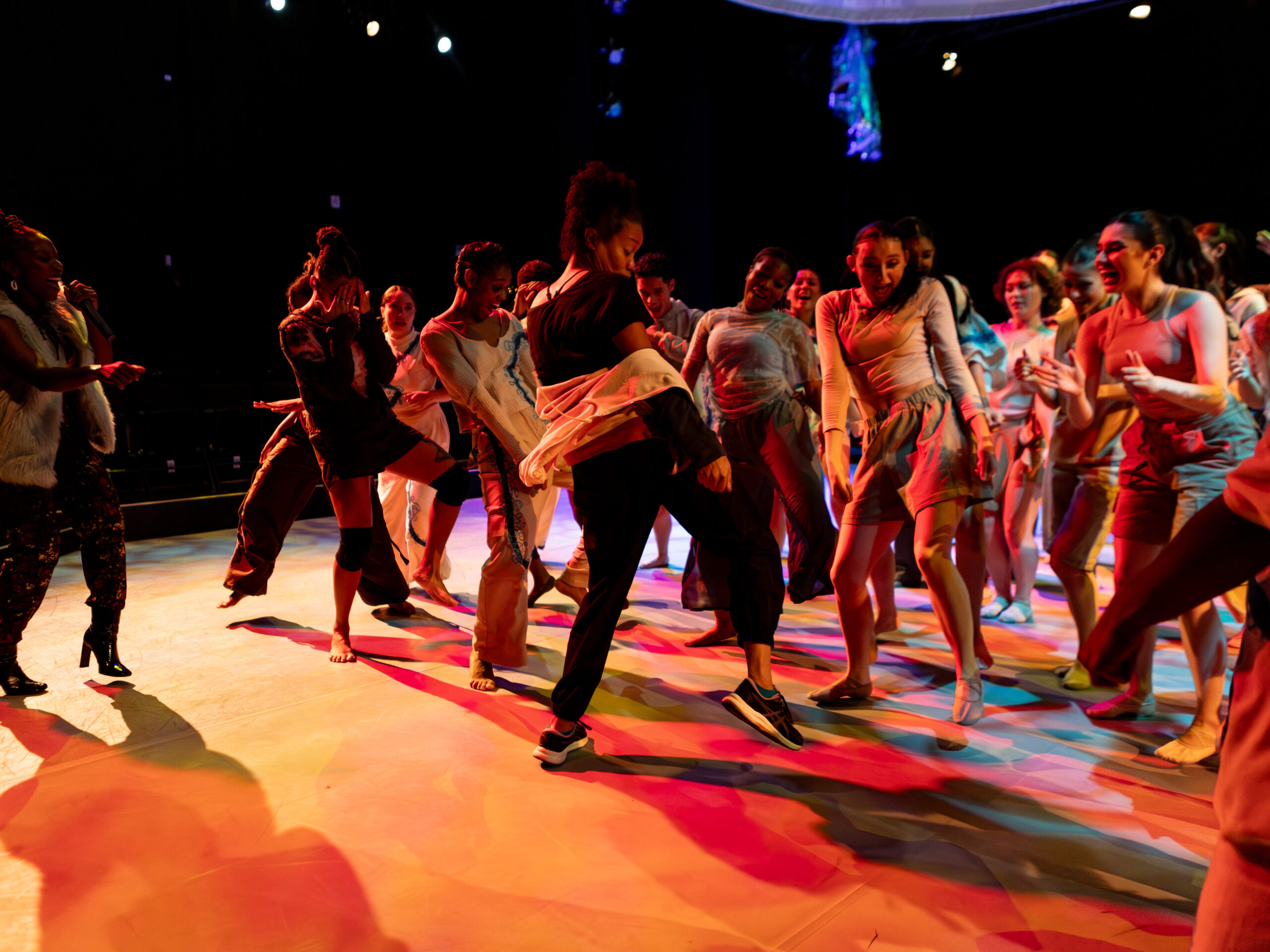 A large group of dancers dance joyfully together on stage, with an MC speaking into a mic behind them