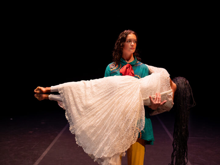 A dancer in a colorful turquoise shirt holds another dancer wearing a white dress, who is in a plank position parallel to the ground