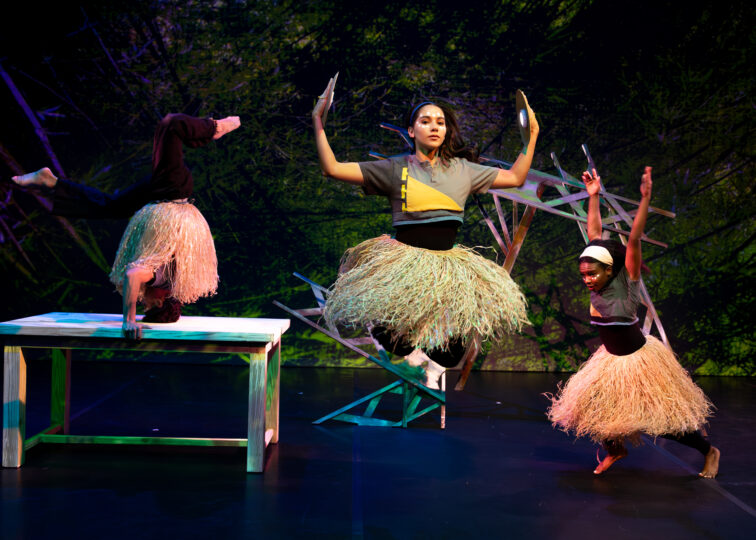 Three dancers perform, one jumping up while holding gold plates in their hands, one in a headstand and one lunging forward with their arms up