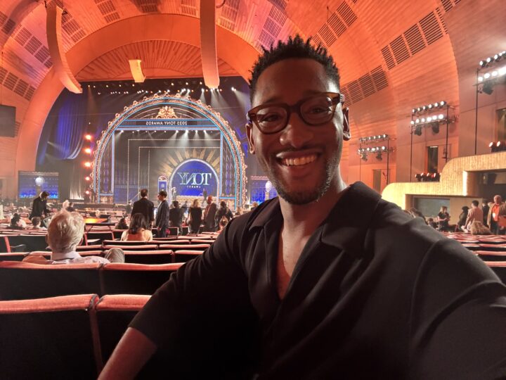 photo of a man with glasses and a black shirt smiling with the seats and stage of the tony awards in the background