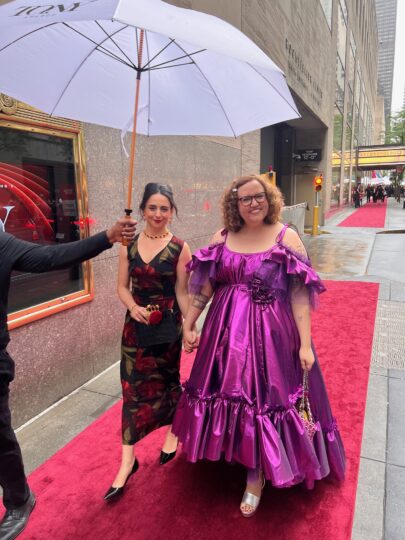 photo of two women holding hands and smiling walking down red carpet in formal gowns
