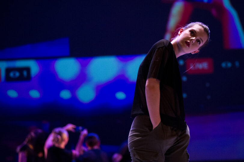 a dancer looks forward with her head tilted to the side and her hand in her pocket, with livestreamed performers projected in the background