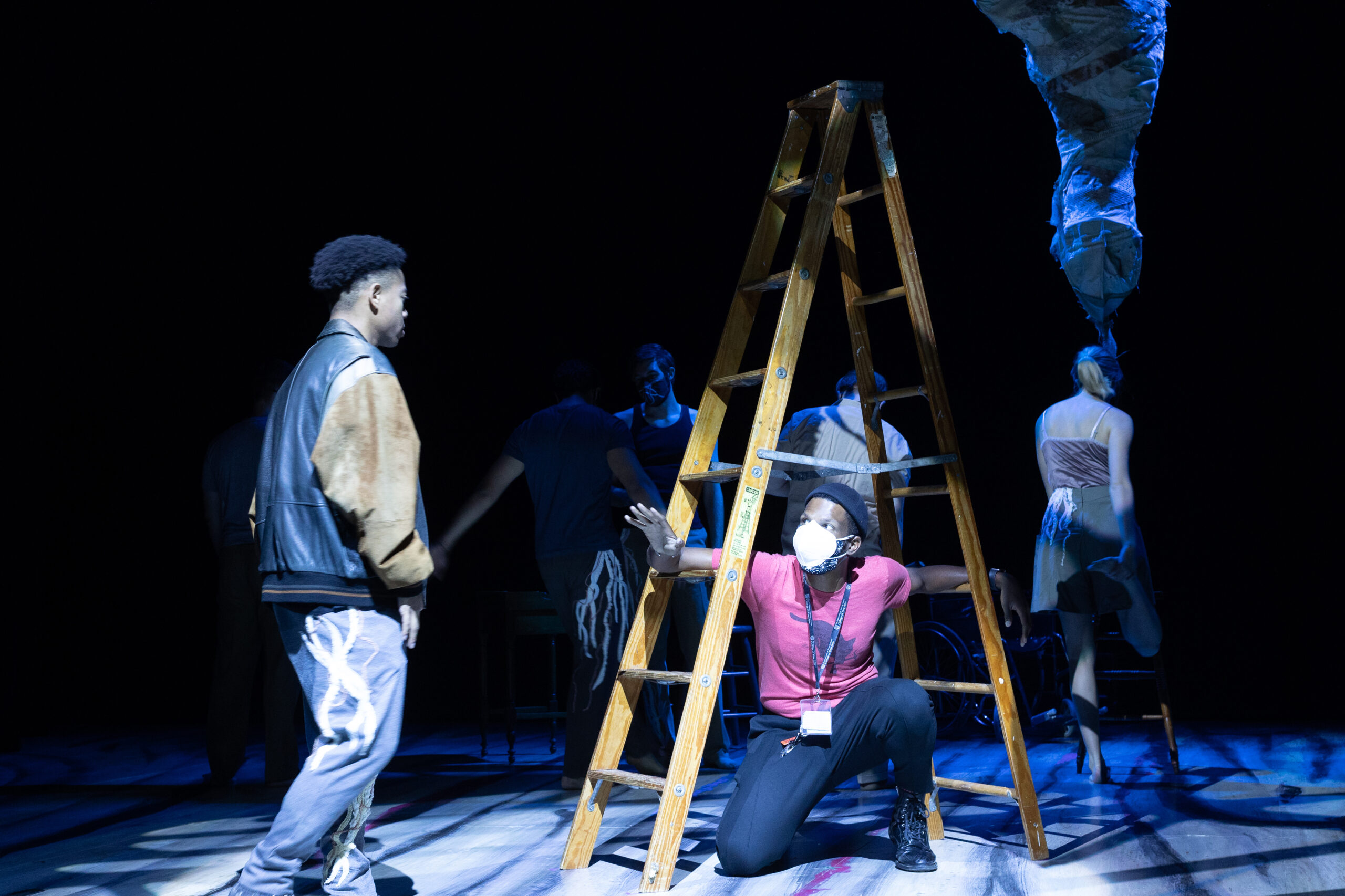 A director sits under a ladder demonstrating a post to an actor who watches
