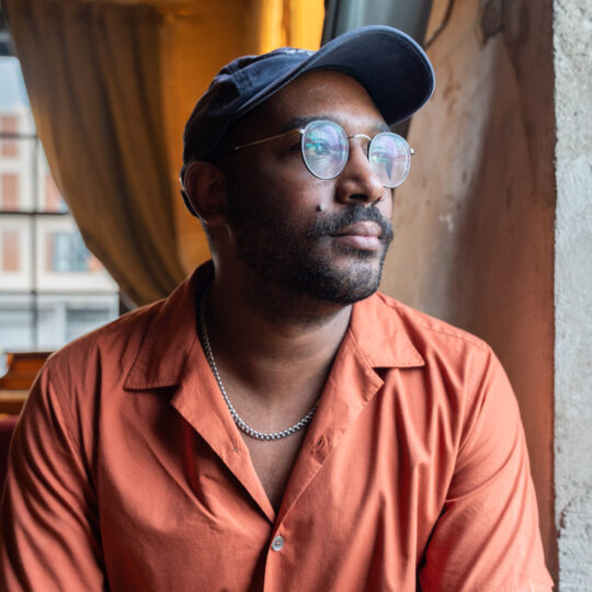 artist with round glasses, a baseball cap and an orange shirt
