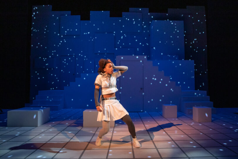 an actress with a shocked expression holds her hand to her head, with blue projections behind her