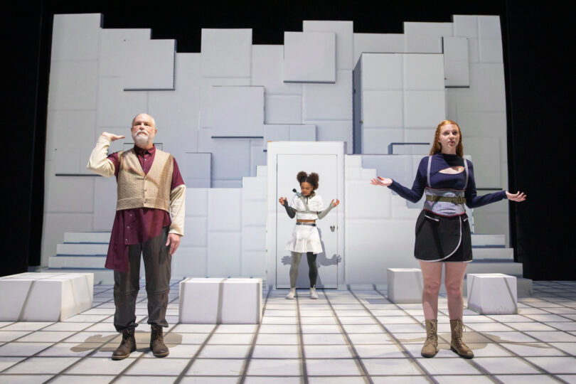 three actors stand on stage; one holds his hand up as if balancing an imaginary tray, while the other two hold their arms up in a shrug