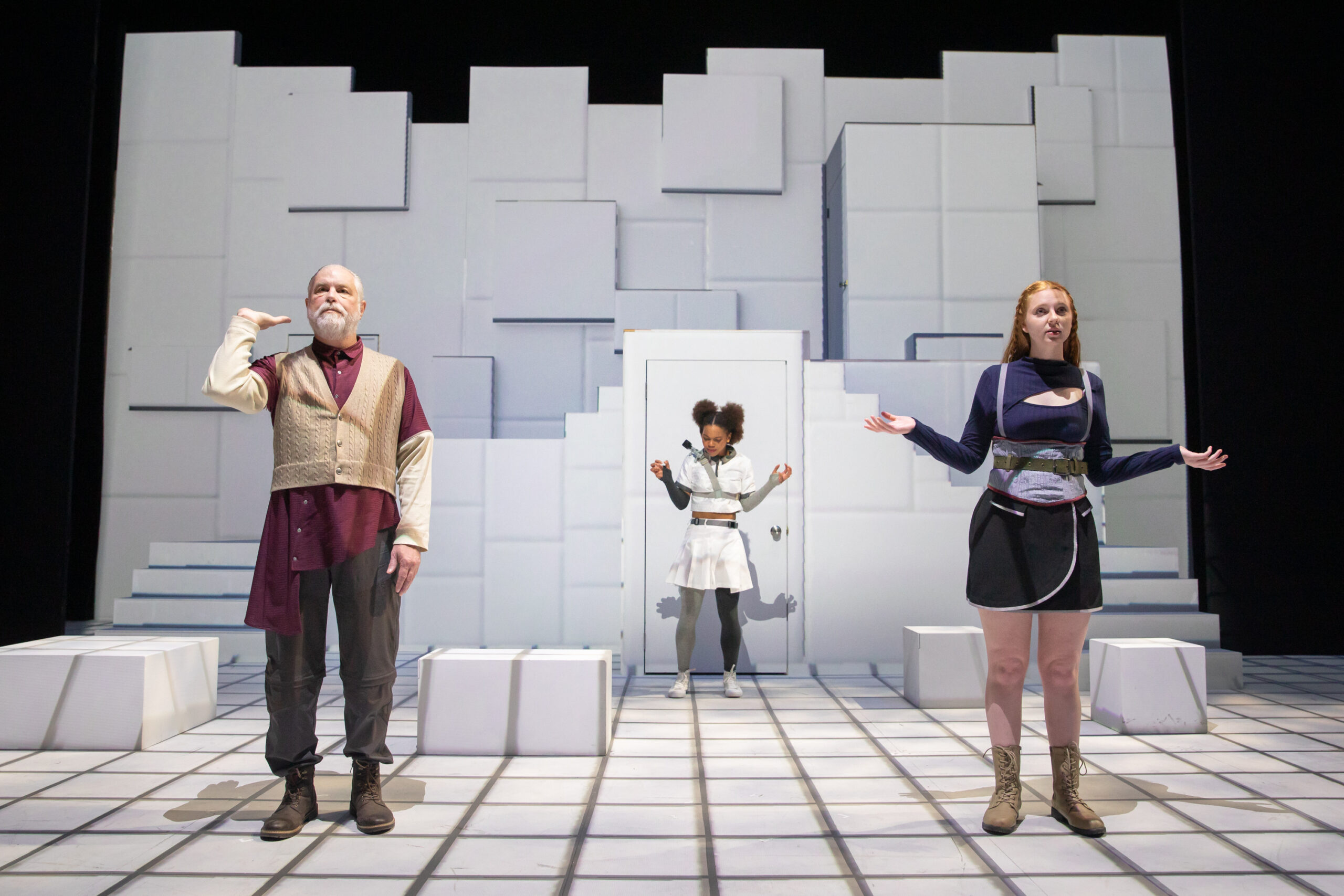 three actors stand on stage; one holds his hand up as if balancing an imaginary tray, while the other two hold their arms up in a shrug