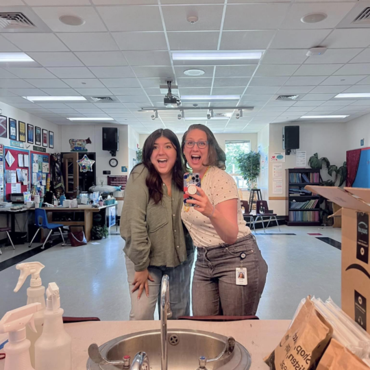 Morgan Co and Briana Bower take a photo in the mirror with their classroom behind them, both looking very excited