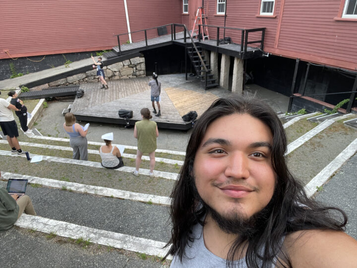 Simon Salinas takes a selfie, with folks rehearsing on an outdoor stage behind him