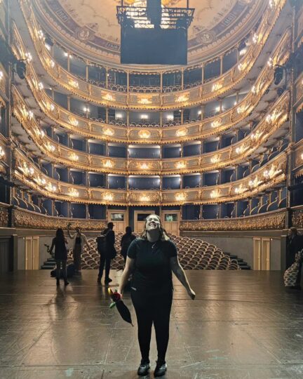 Alex Titsworth standing excitedly on a large stage with an ornate, multi-floor theatre house behind them