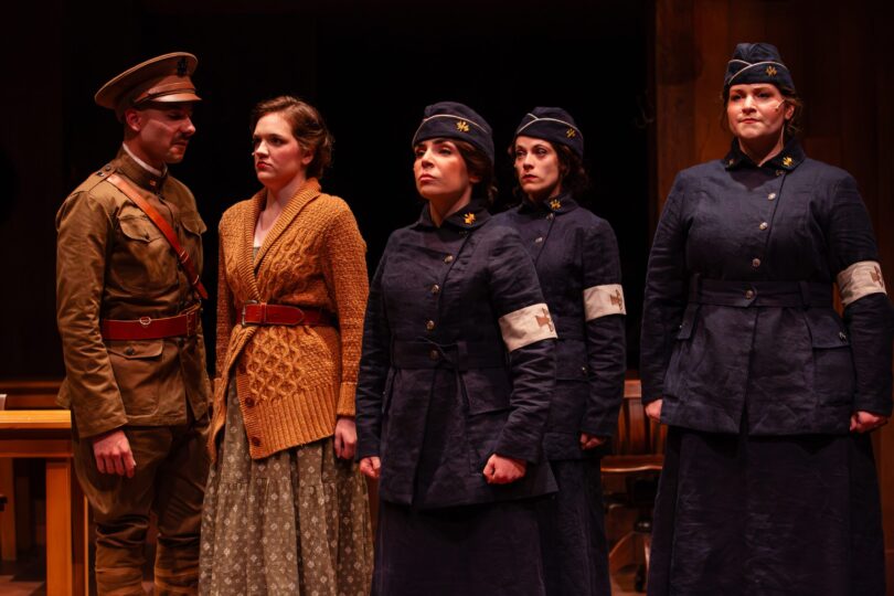 A scene from THE HELLO GIRLS in which three women in dark uniforms stand at attention while a fourth woman in a sweater and a man in a uniform stand next to them
