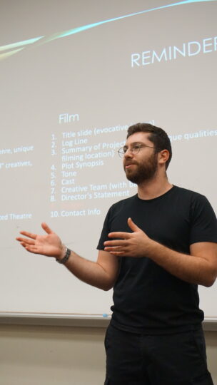 David Treatman stands in front of a projector screen with instructions on it, facing a classroom of students