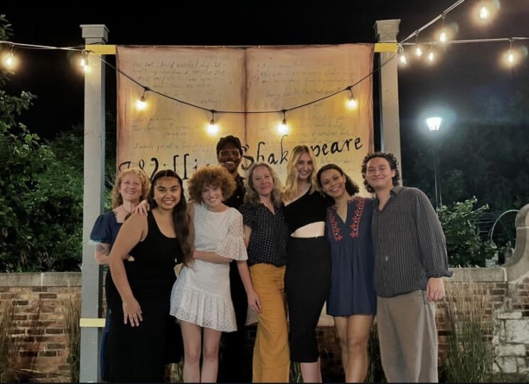 Eight students, alumni, faculty and former faculty pose together in front of a banner that says WILLIAM SHAKESPEARE