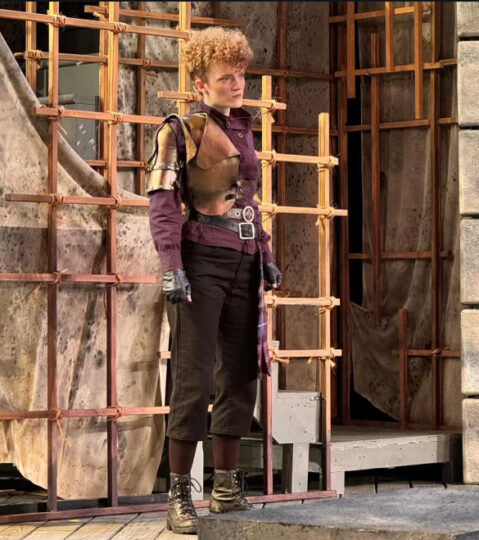 Kira Small performs in a scene from MACBETH, standing and staring forward with intensity.