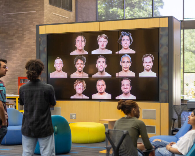 Photo of facepiece from the cohen new works festival. it shows 11 faces on a massive tv screen in the fine arts library. 
