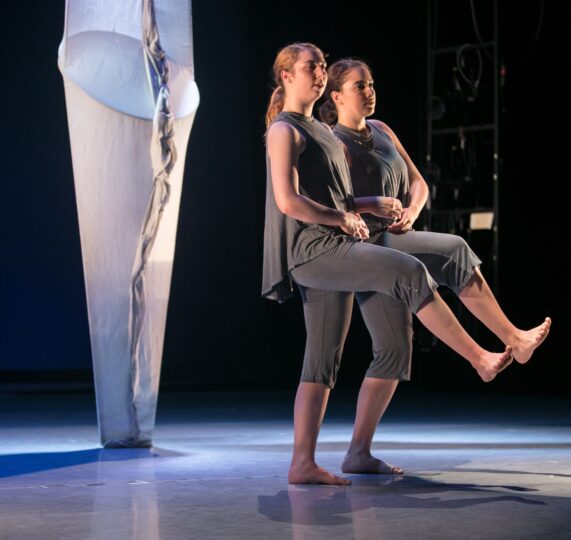 Photo of two dancers in grey moving in tandem with a white fabric vertical set piece behind them.
