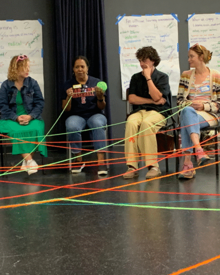 Woman holding a bright green ball of yarn that has been strung across a circle of teachers