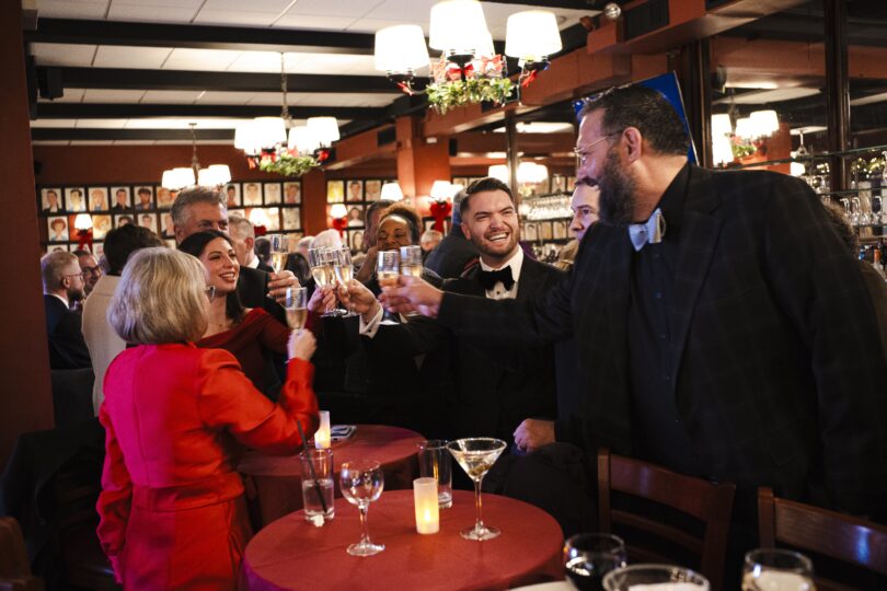 photo of a group of people in tuxedos and formal attire clinking champagne glasses together and smiling