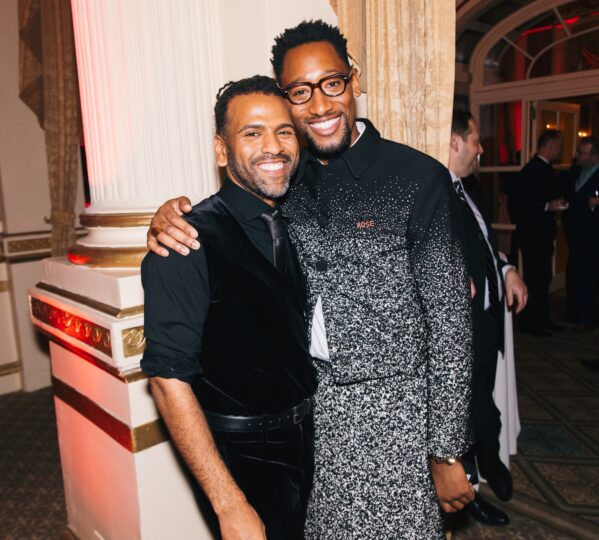 photo of two men in black, formal clothes with arms around each other smiling