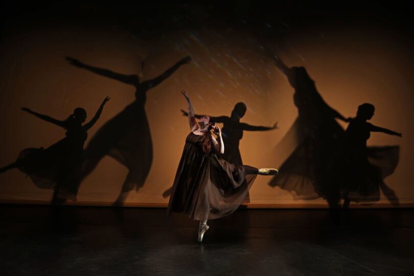 One dancers balances in an arabesque en pointe, with the shadows of four other dancers behind her