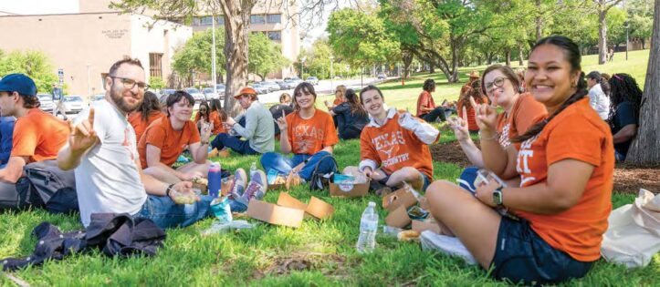 UT Social Work students in burnt orange shirts relaxing on the grass outdoors on the UT campus.
