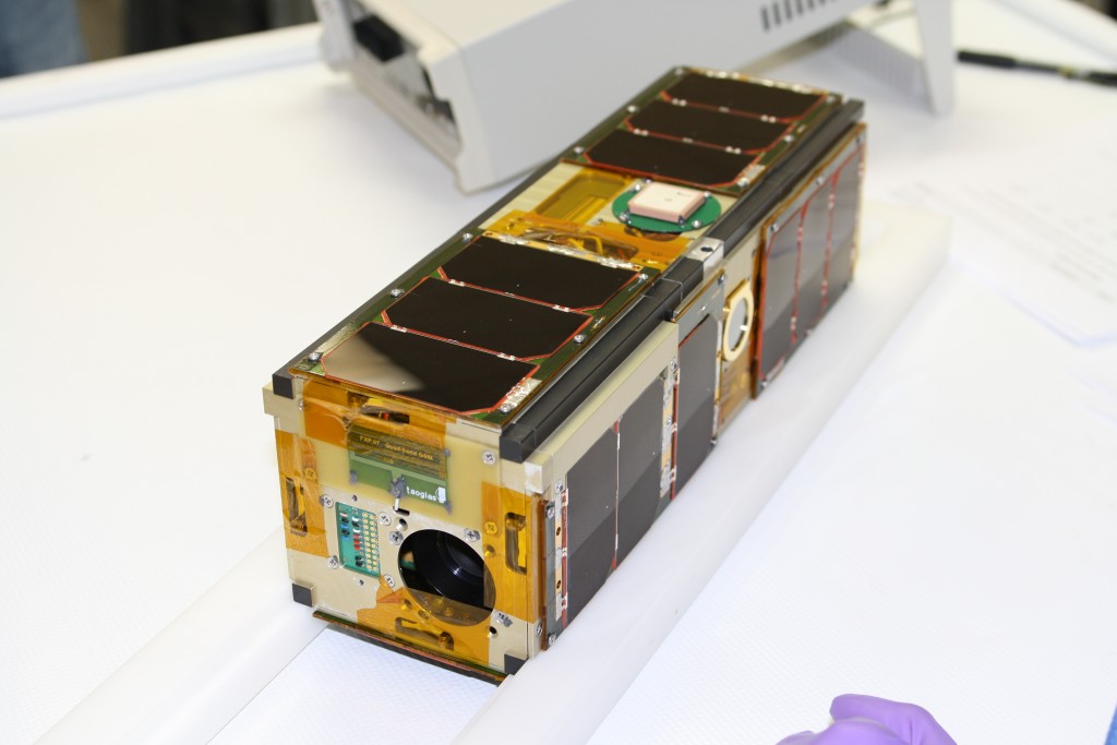 A student-built CubeSat developed by the Texas Spacecraft Laboratory, displayed on a white surface in a research lab.”