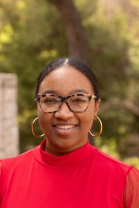 A headshot of black woman with shell glasses and pulled back hair. She wears golden hoops and a red blouse. 