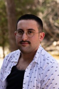 A headshot a caucasian  man with a buzzed black hair and a mustache. Wearing a white button down shirt over a black t-shirt 