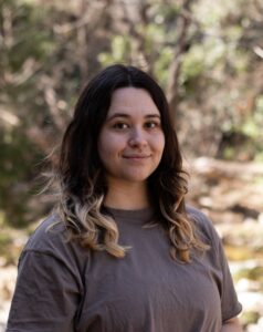 A headshot of a Caucasian person in a gray T shirt with highlights in their long wavy hair. 