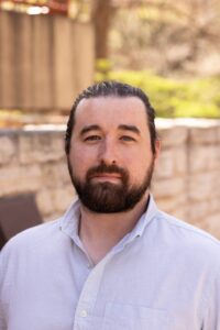 A headshot of a caucasian man in with a dark beard and pulled back hair. He is in a cool white button down. 