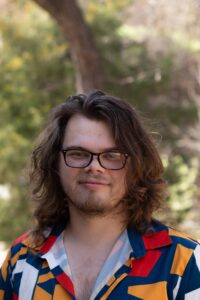 A headshot of a caucasian person with long fluffy hair and a small beard  wearing shell glasses and a bright multicolored button down shirt with Blue, Yellow and Red shapes on it. 