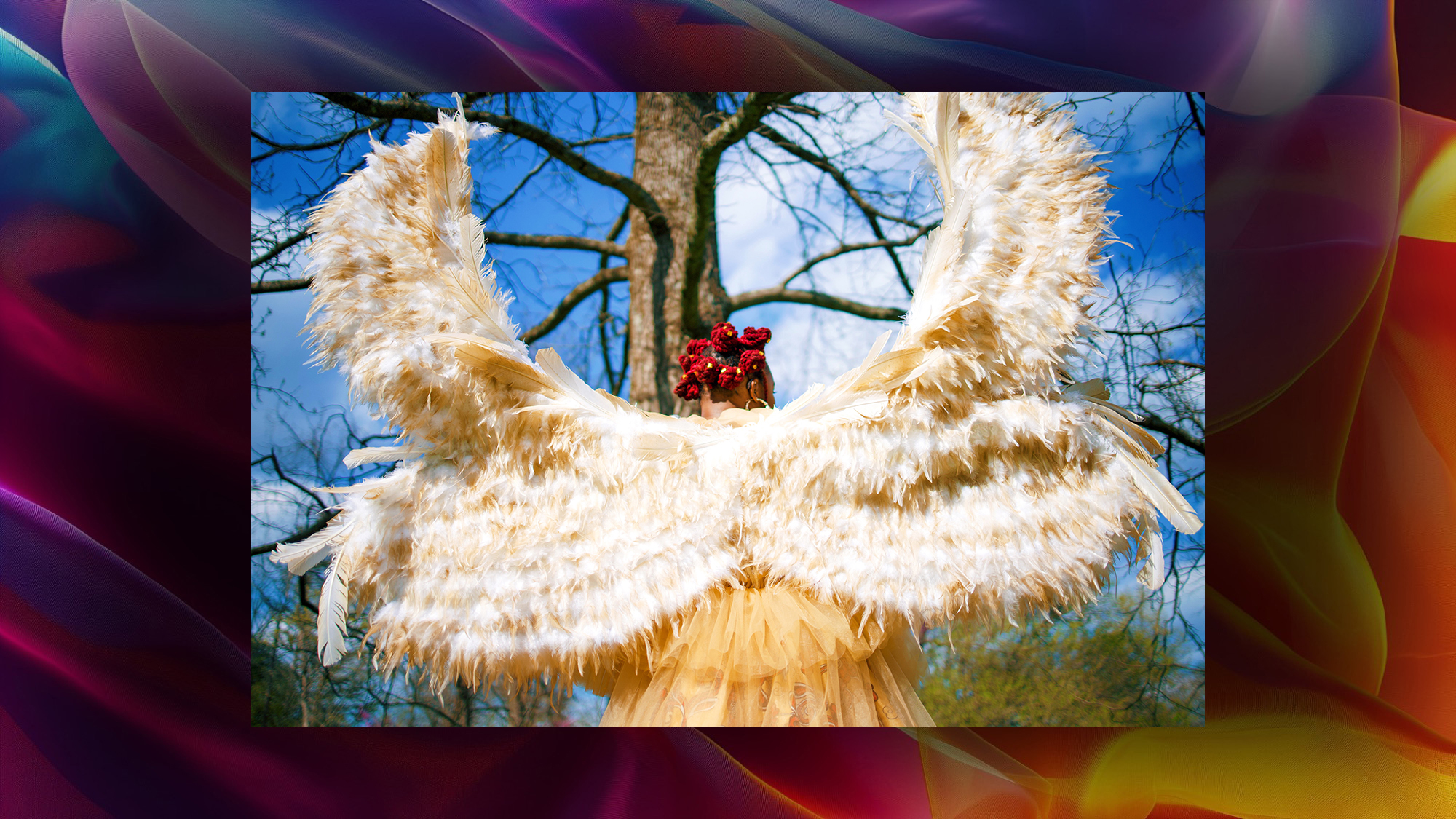 A colorful field with single image on it. The picture is black woman in a costume of an angel with four wings. Her hair is red and pulled back into tightly twisted buns. The wings are cream and white in color and she wears an ivory sheer robe over African wax printed fabric dress. 