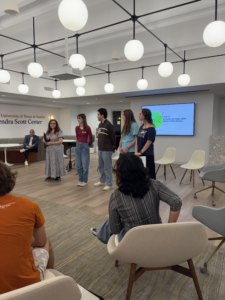 Roxanne Schroeder-Arce, Associate Dean of UTeach Fine Arts standing with Student Advisory Council (UFASAC) students in the Kendra Scott Center making announcements about the panel guests. On the screen behind them is a slide that states"welcome to the Future of Fine Arts Education panel"