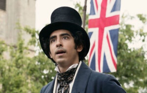 Movie still of Ango-Indian actor Dev Patel as David Copperfield, wearing a tall hat standing in front of a Union Jack